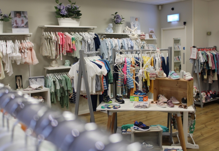 The inside of a boutique childrenswear store with a number of rails and shelves filled with children's clothes and accessories from a spring collection.