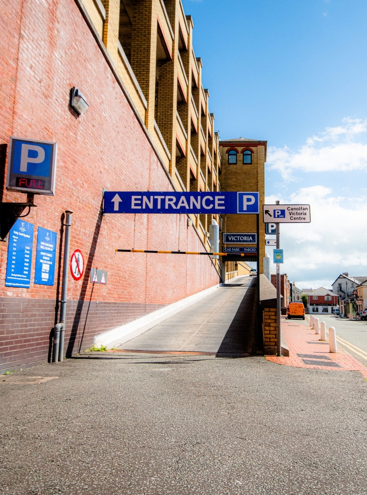 The entrance way to the shopping centre's car park. The shot is taken looking up the ramp which goes up to the first floor of the car park. It is a sunny day and the sky is blue.