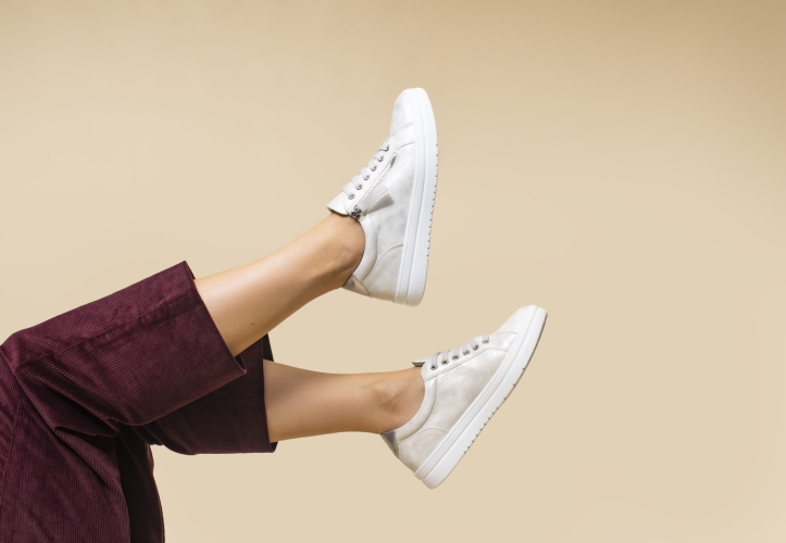 A pale gold background with legs sticking up into the shot wearing Pavers white trainers and cropped burgundy trousers.